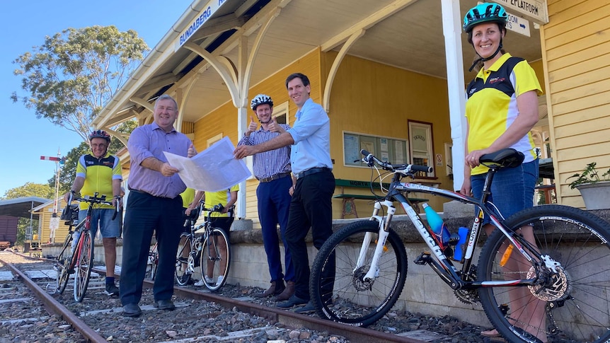 Three politicians stand on railway tracks in front of an old platform, holding a map and thumbs up, flanked by cyclists