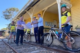 Three politicians stand on railway tracks in front of an old platform, holding a map and thumbs up, flanked by cyclists