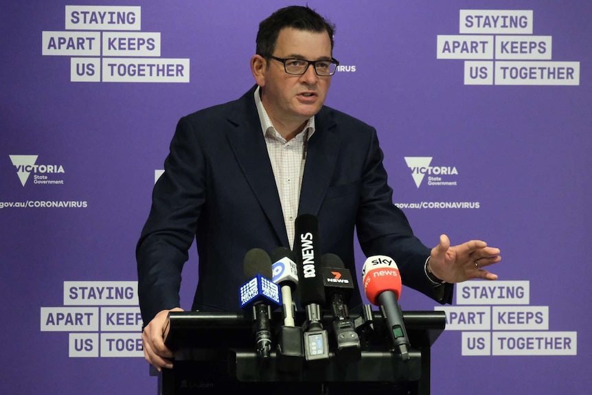 Victorian Premier Daniel Andrews standing at a lectern in front of microphones with a purple backdrop.