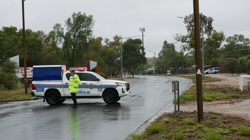 A police man and his police van parked across a road with police tape, in the rain.