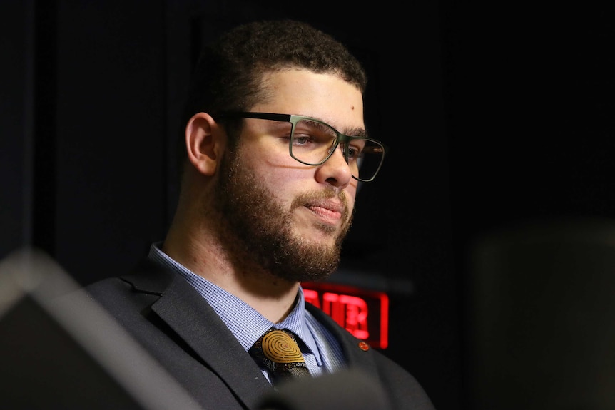 A young, bearded and bespectacled man sits in what appears to be a television studio.