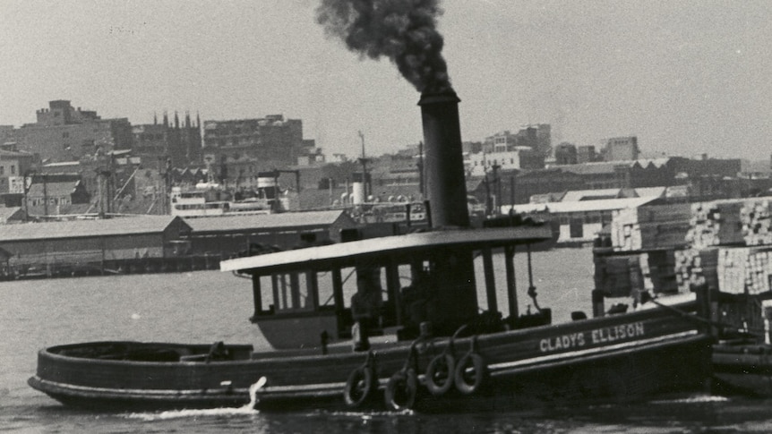 A black and while photo of a timber steam tugboat, tied up at a dock. Steam coming from its stack.