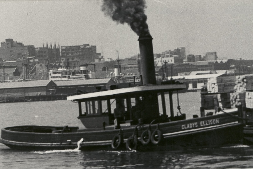 A black and while photo of a timber steam tugboat, tied up at a dock...steam coming from its stack . 