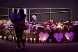 A mother holds her young daughter as they look at a memorial with flowers, candles and signs.
