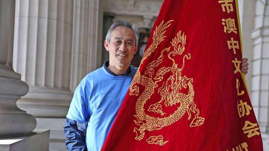 A man with greying hair smiles as he holds a large red flag with a golden dragon embroidered on it.