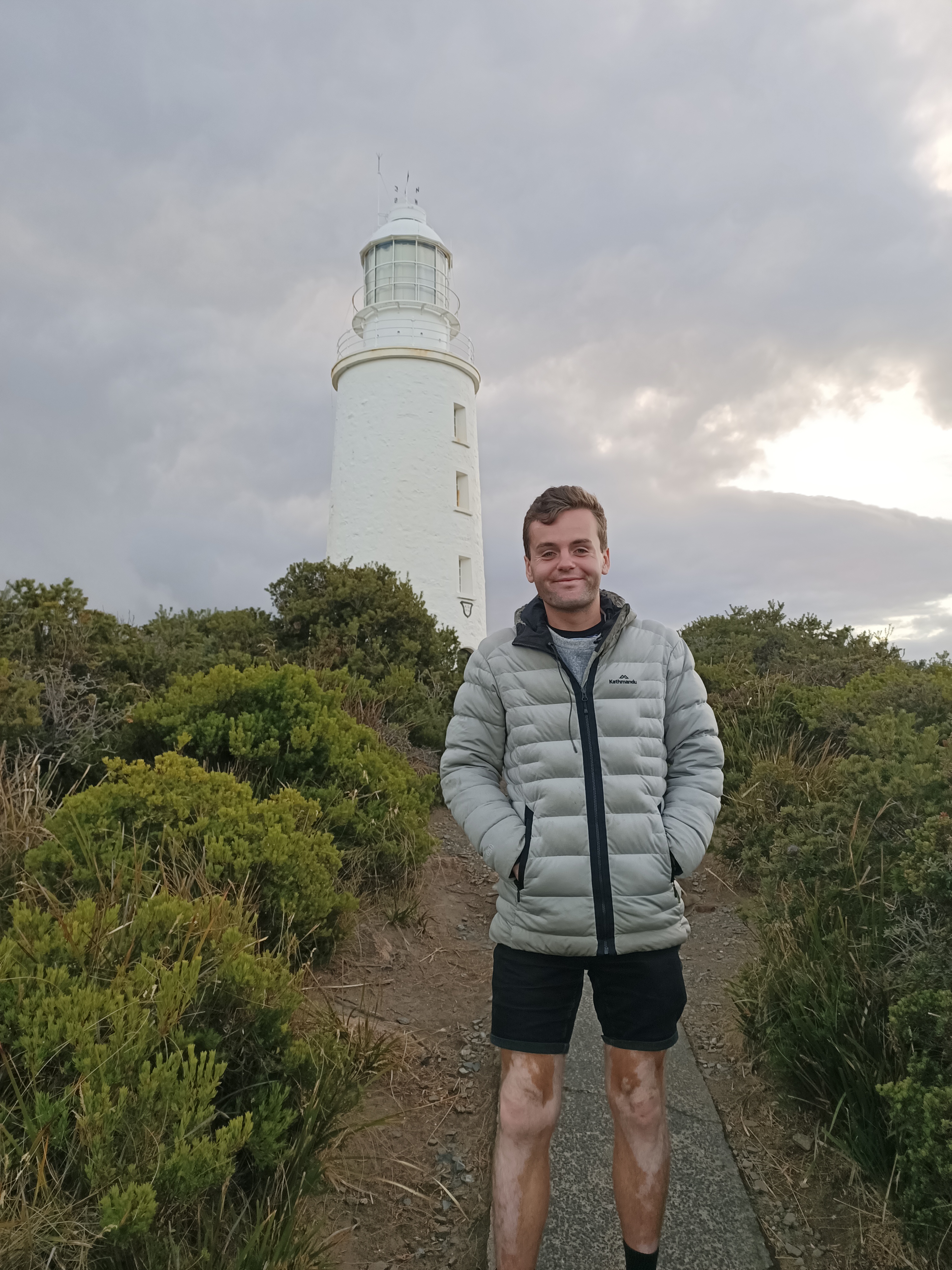 A man wearing shorts and a hoodie smiles in front of a white lighthouse.