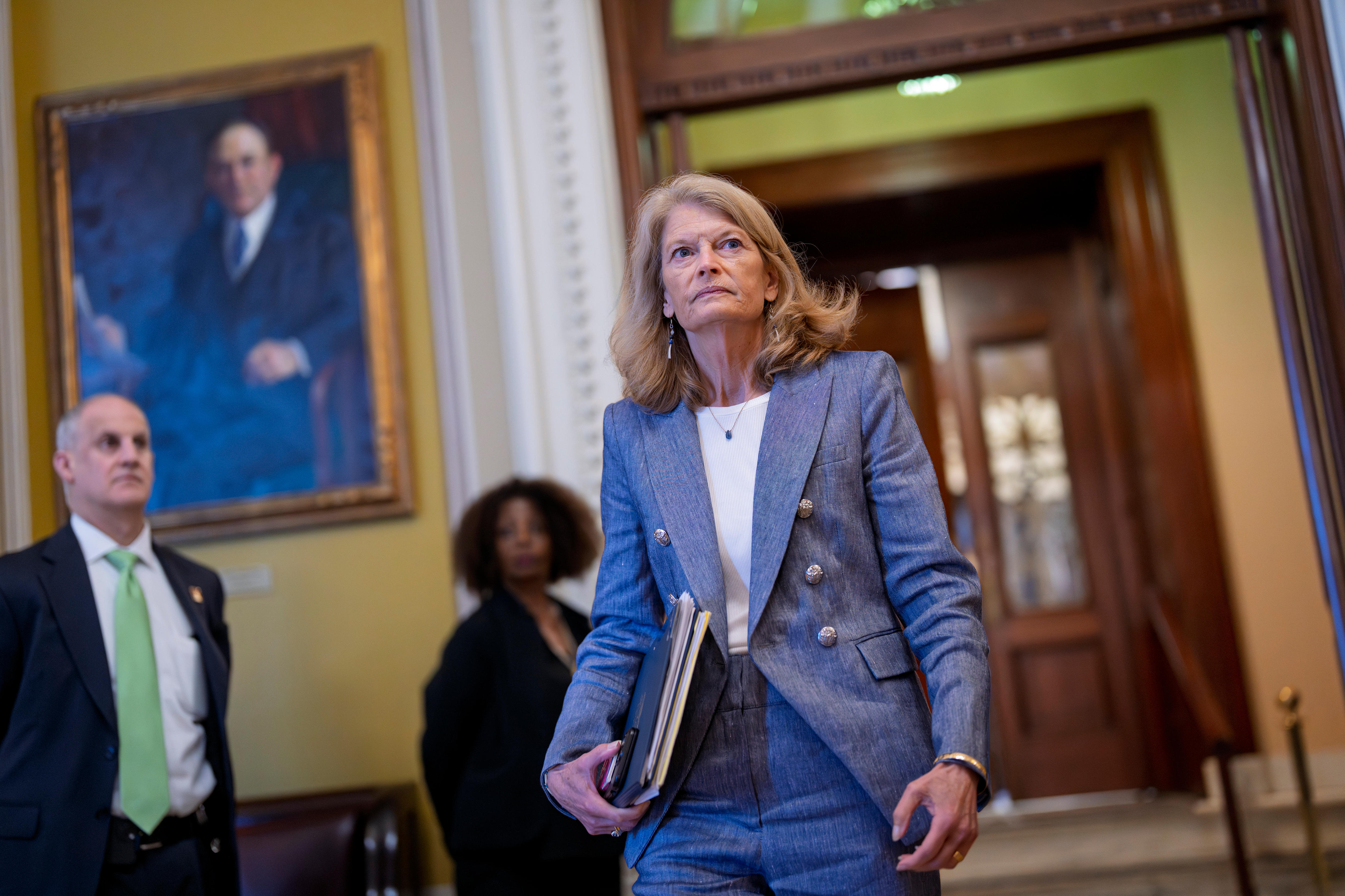 A woman in a grey pantsuit holds a binder to her hip as she walks through a hallway