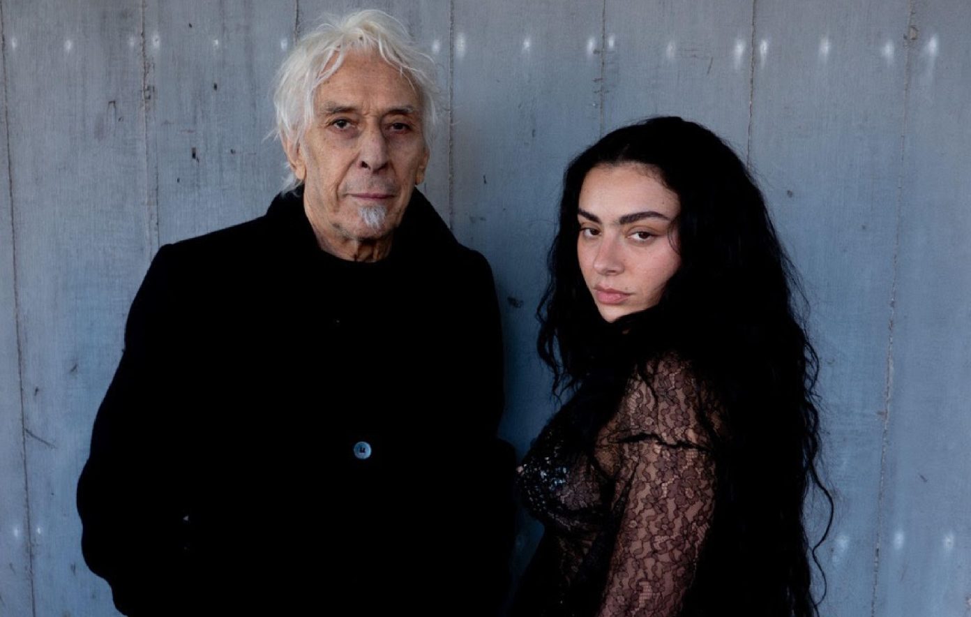 An ashen-haired John Cale poses in a black trenchcoat with Charli XCX, wearing black lace sleeved top