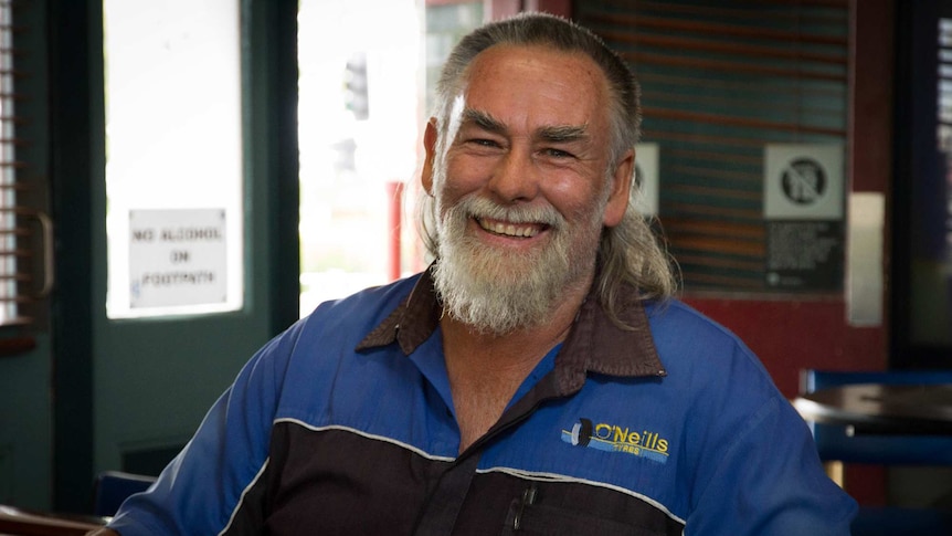 A man with a beard, a mullet hairstyle, and a mechanics shirt, sits smiling in a country pub.