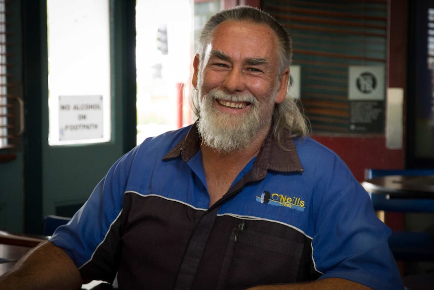 A man with a beard, a mullet hairstyle, and a mechanics shirt, sits smiling in a country pub.