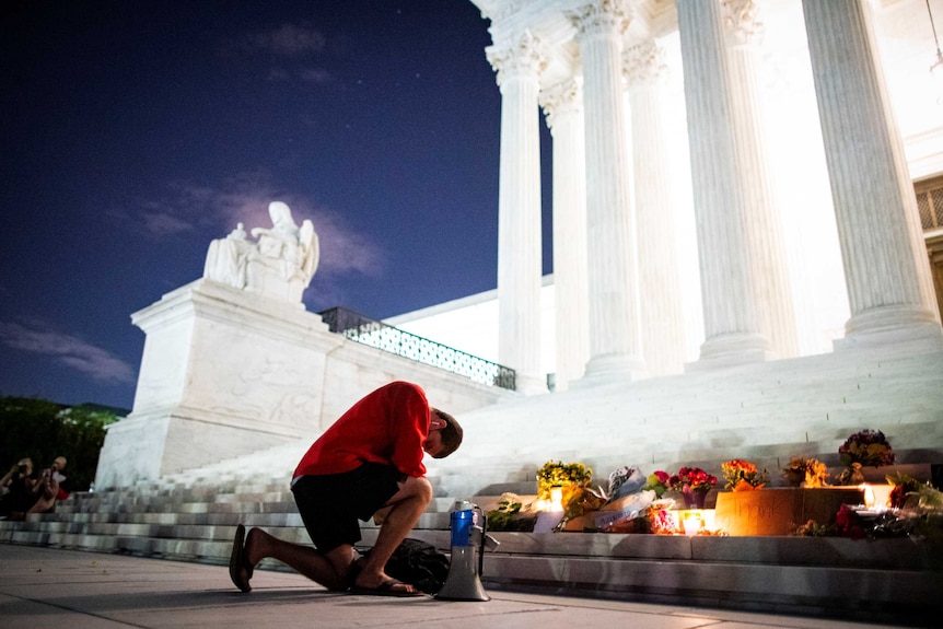 A man kneels in front of a shrine of flowers and candles on the steps of the US Supreme Court
