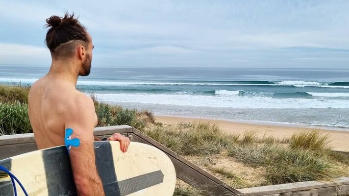 Man holding surfboard looking at the water with a blue insulin patch in his right arm 