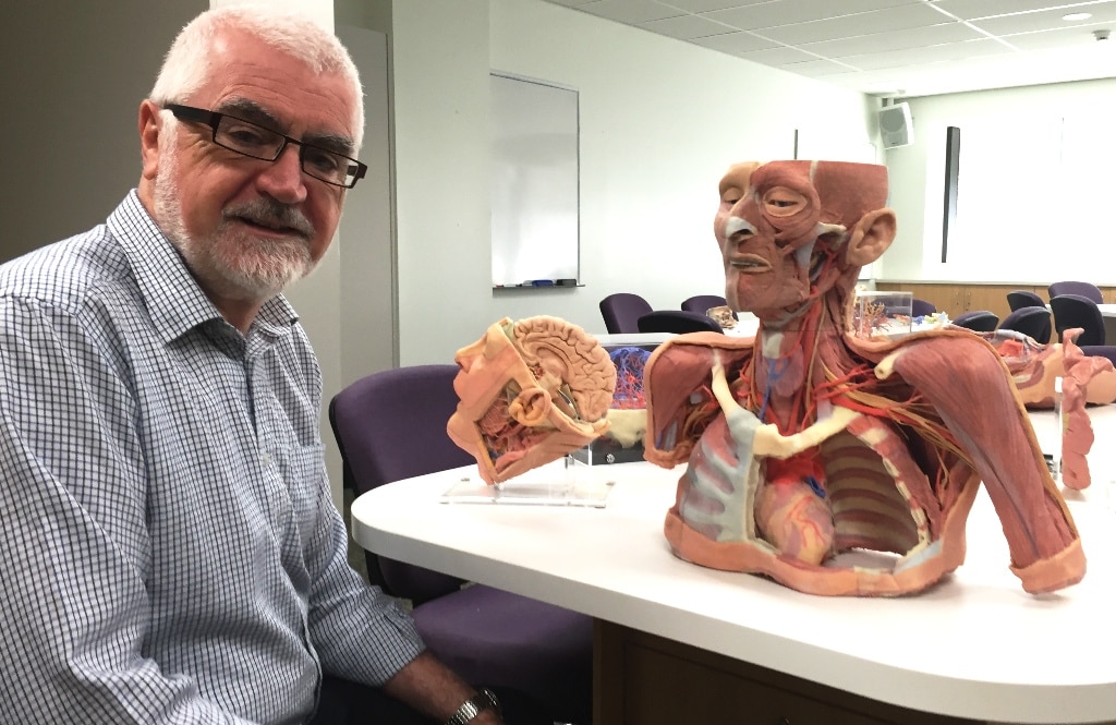 A man with white hair and a check shirt sitting next to a table with replica body parts.