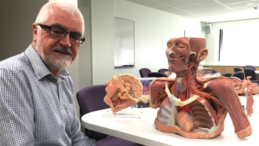 A man with white hair and a check shirt sitting next to a table with replica body parts.