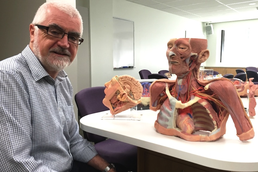 A man with white hair and a check shirt sitting next to a table with replica body parts.