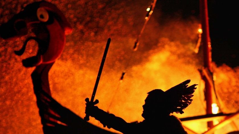 A man dressed as a viking is silhouetted against a burning Viking long ship