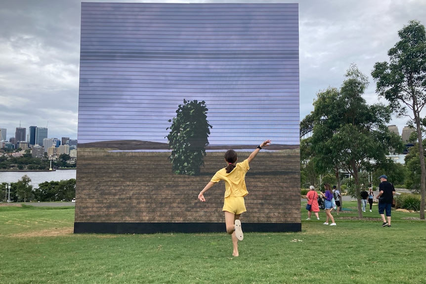 Girl dressed in yellow dances in front of large square screen with leaf-covered man installed on lawn, cityscape in background.