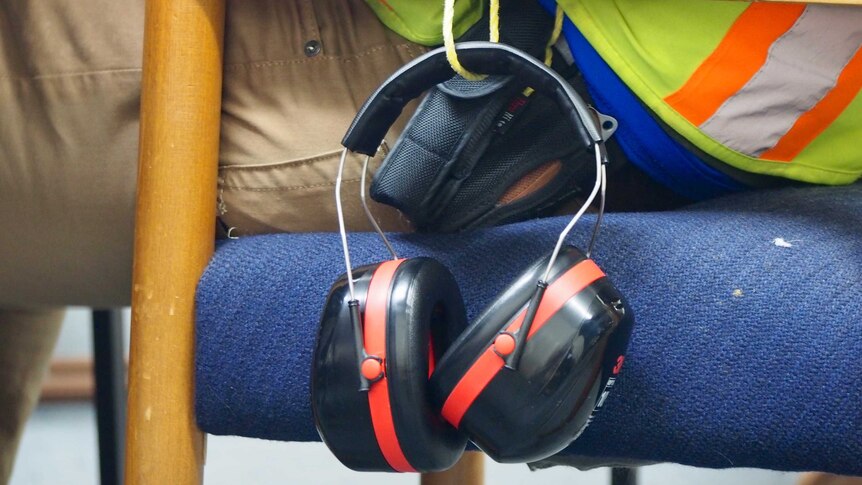 Ear muffs dangle from the side of a chair. The person sitting in it is wearing a high-vis vest.