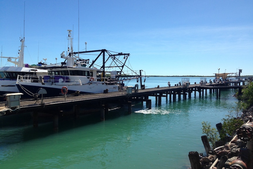 Fishing trawler boat docked next to a long pier off the coast line on a bright sunny day. 