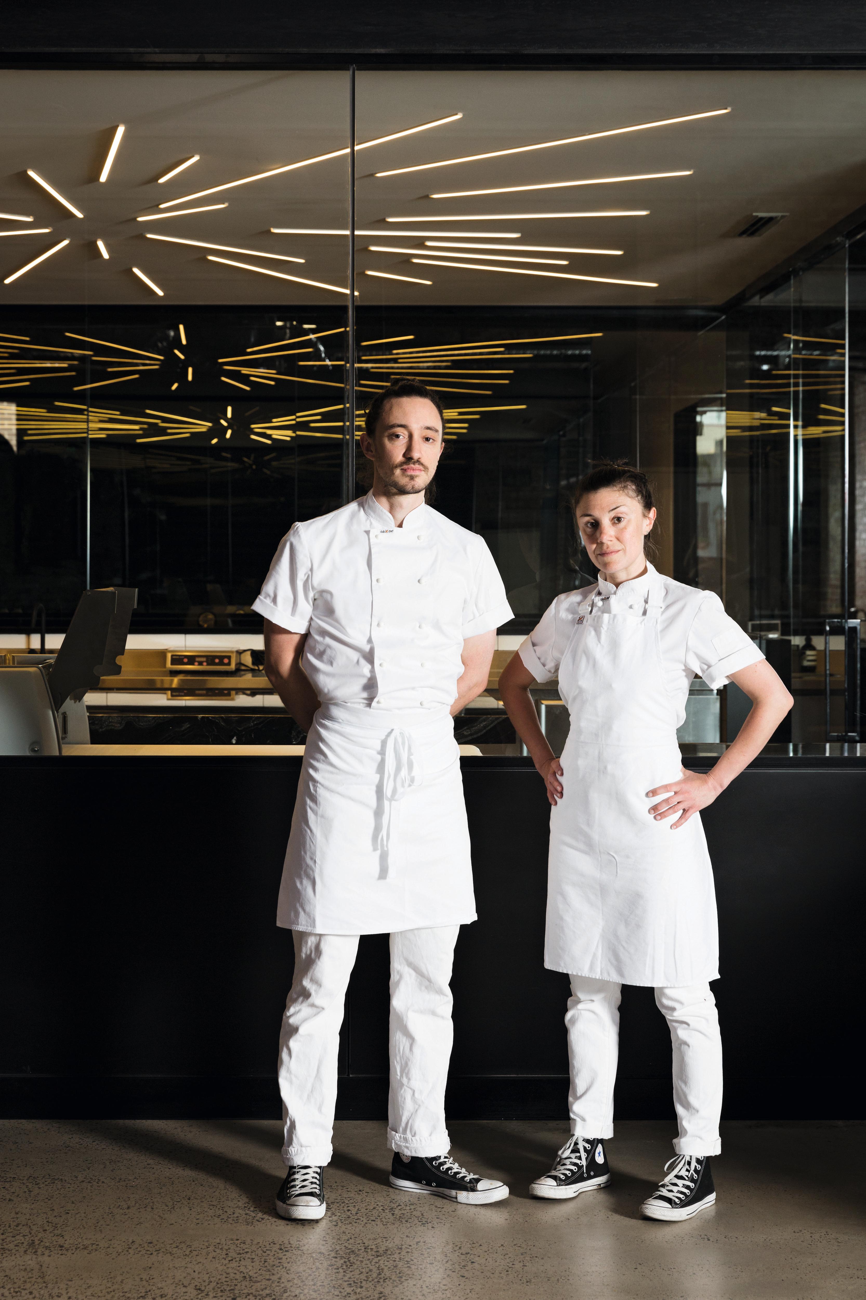A young man and woman in white chef clothes standing in a dark room, equipment in background
