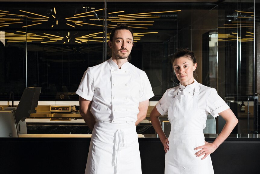 A young man and woman in white chef clothes standing in a dark room, equipment in background