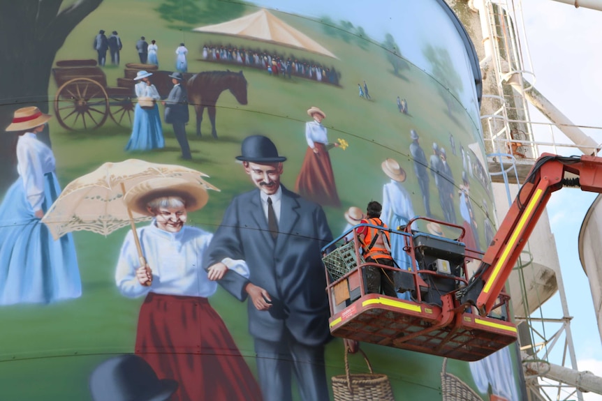 close up of man in high vis on cherry picker painting silo artwork of old picnic.