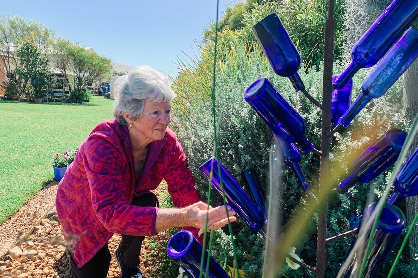 a woman kneels down in a garden bed inspecting a sculpture made from blue glass bottles