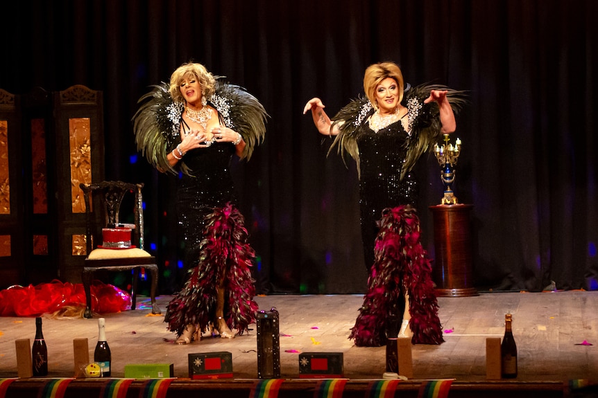 Two drag queens stand on a stage wearing matching costumes with large feather capelet an floor-length sequin gowns