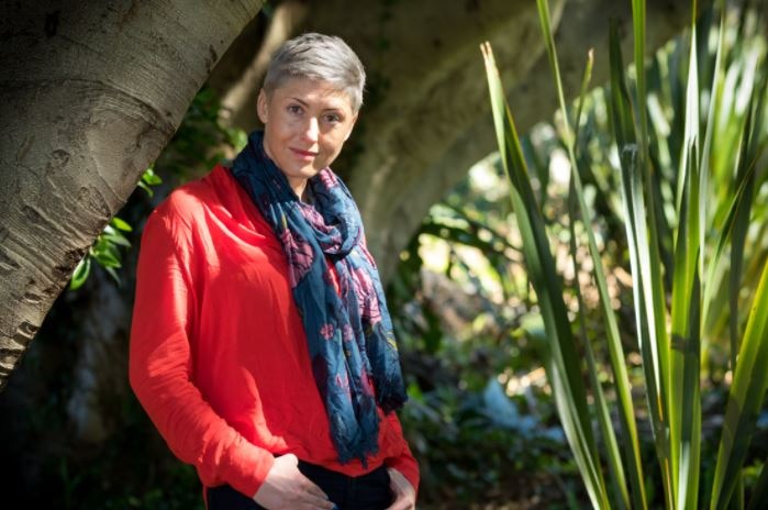 A woman with short blonde hair, wearing a red top and colourful scarf stands in a garden.