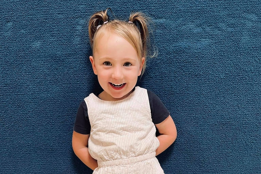 A young girl smiles while standing against a blue wall indoors looking at the camera, wearing a cream jumpsuit and dark shirt.