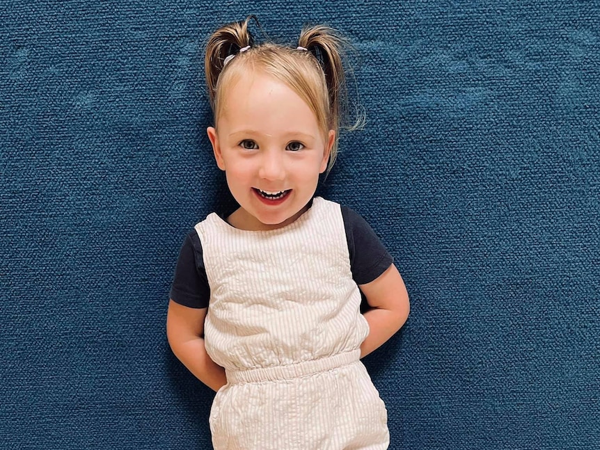 A young girl smiles while standing against a blue wall indoors looking at the camera, wearing a cream jumpsuit and dark shirt.