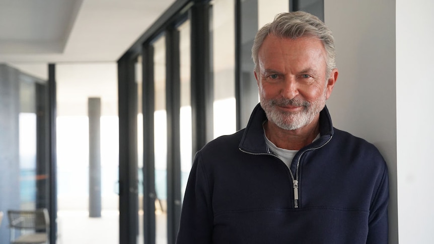 Sam Neill leaning against a wall smiling, glass doors behind him.