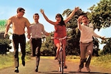 The four band members of The Seekers run and cycle down a sunlit path sided by trees and greenery.