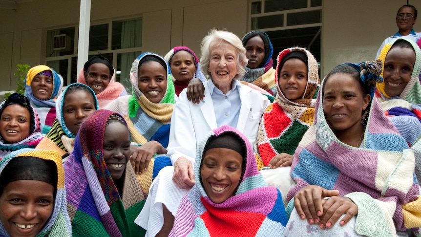 An older woman wearing a white doctor's coat stands with a group of smiling women wearing colourful blankets