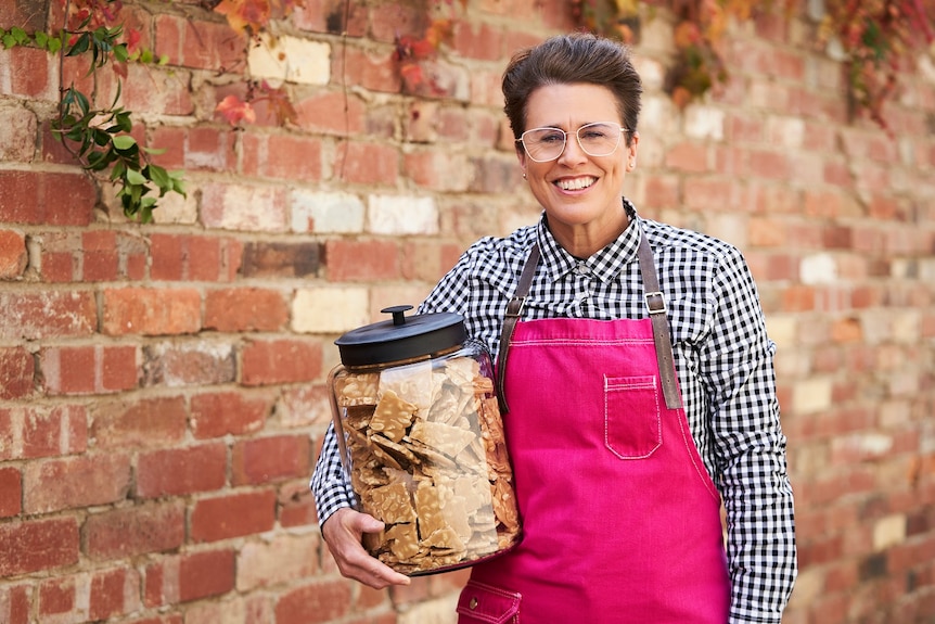 Greta Donaldson wearing a hot pink apron standing in front of a brick wall, holding a large jar of peanut brittle.