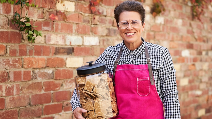 Greta Donaldson wearing a hot pink apron standing in front of a brick wall, holding a large jar of peanut brittle.