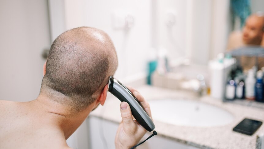 The back of a man's head. He holds clippers and is shaving his hair off in the bathroom. 
