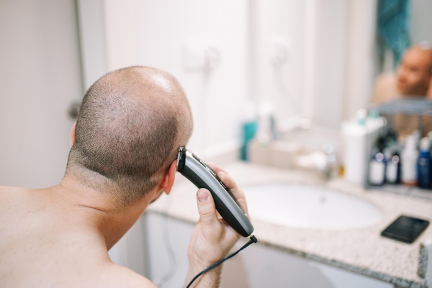 The back of a man's head. He holds clippers and is shaving his hair off in the bathroom. 