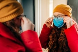 A girl with glasses fits a face mask in front of a mirror