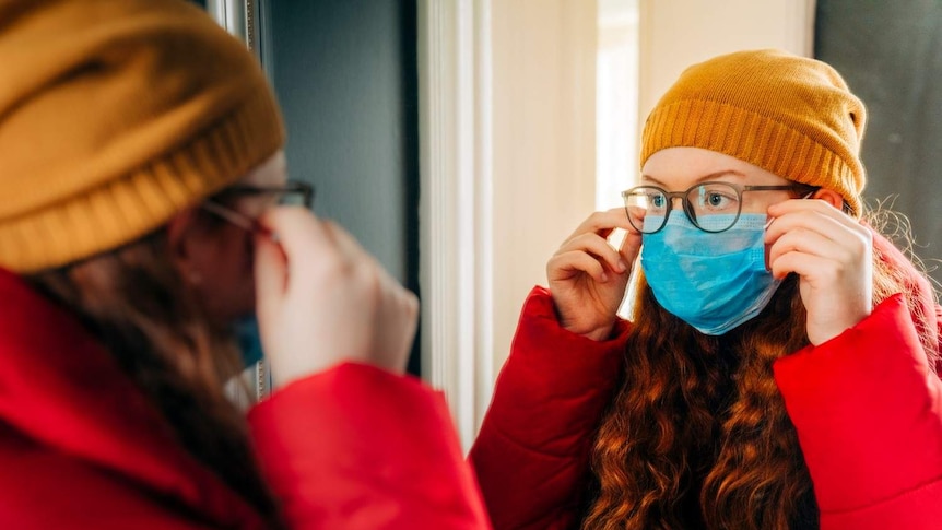 A girl with glasses fits a face mask in front of a mirror