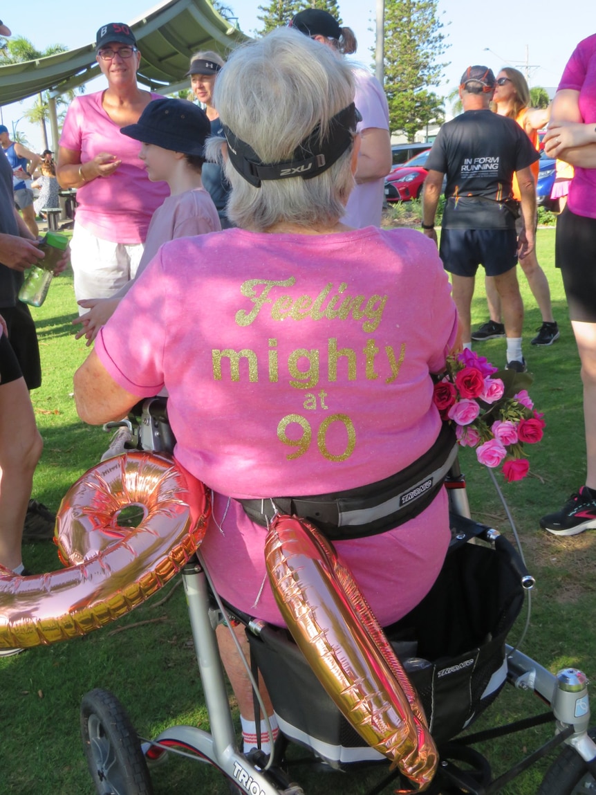 An elderly woman with her back turned sits on her mobility walker with balloons and flowers decorating it