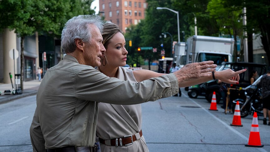 An older man with grey hair and woman in khaki jumpsuit stand close together in the middle of a city street, both pointing.