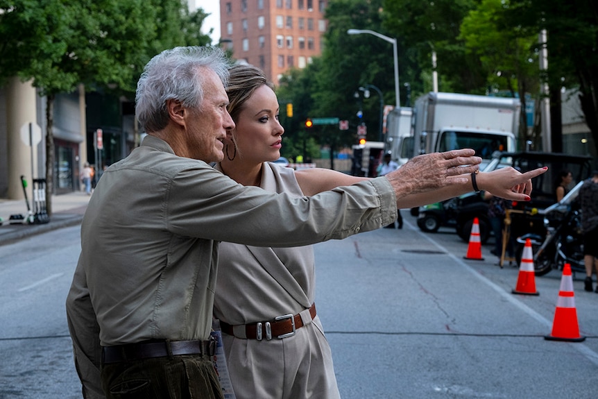 An older man with grey hair and woman in khaki jumpsuit stand close together in the middle of a city street, both pointing.