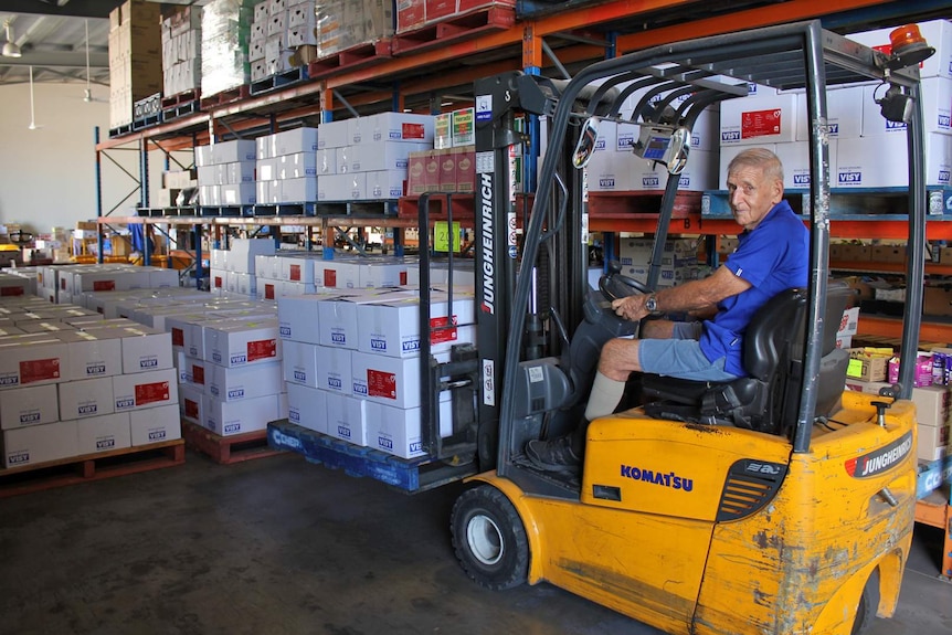 An elderly man drives a forklift with a pallet of boxes on the front.