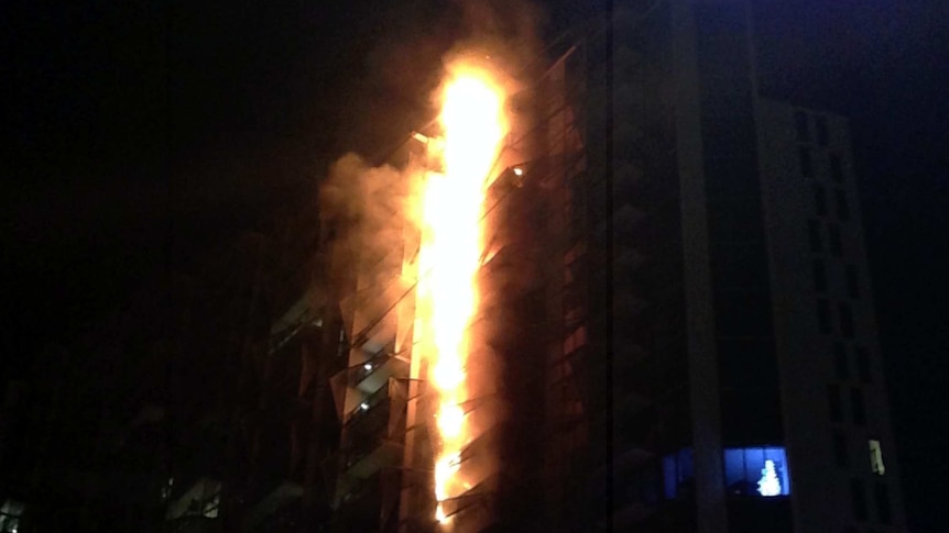 A fire burns up the side of a dark apartment building at night time.