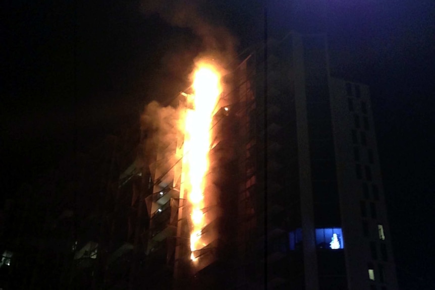 A fire burns up the side of a dark apartment building at night time.