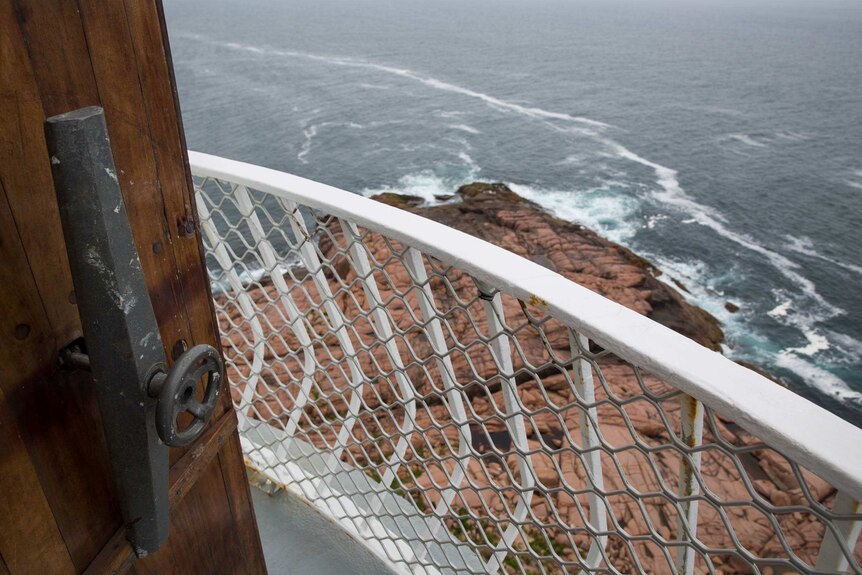 A rocky outcrop and crashing surf is seen beyond a heavy wooden door and white railing at the top of the lighthouse.