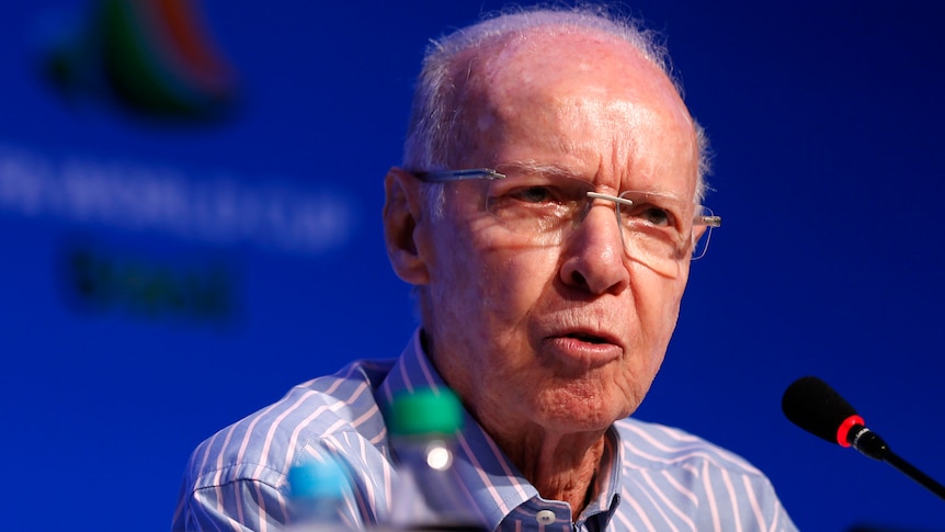 Mário Zagallo wearing glasses speaks at a desk at a press conference