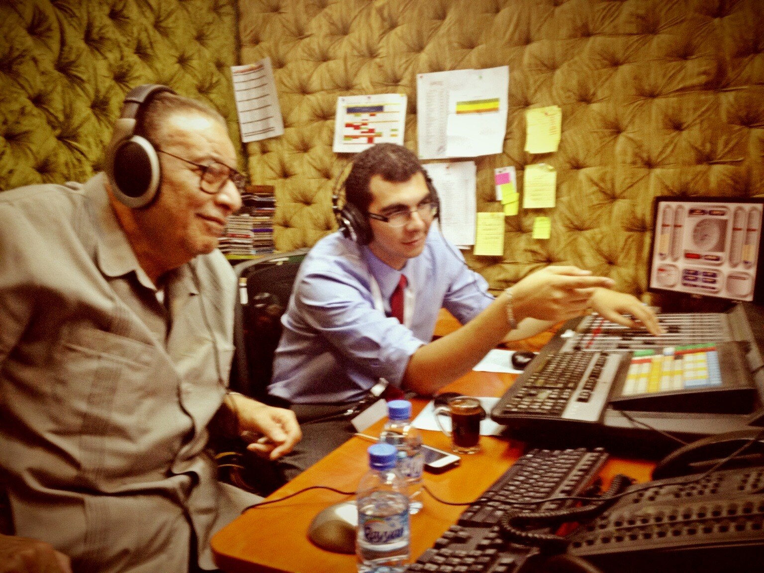 Two men wearing headphones looking at a panel of buttons in a radio studio.