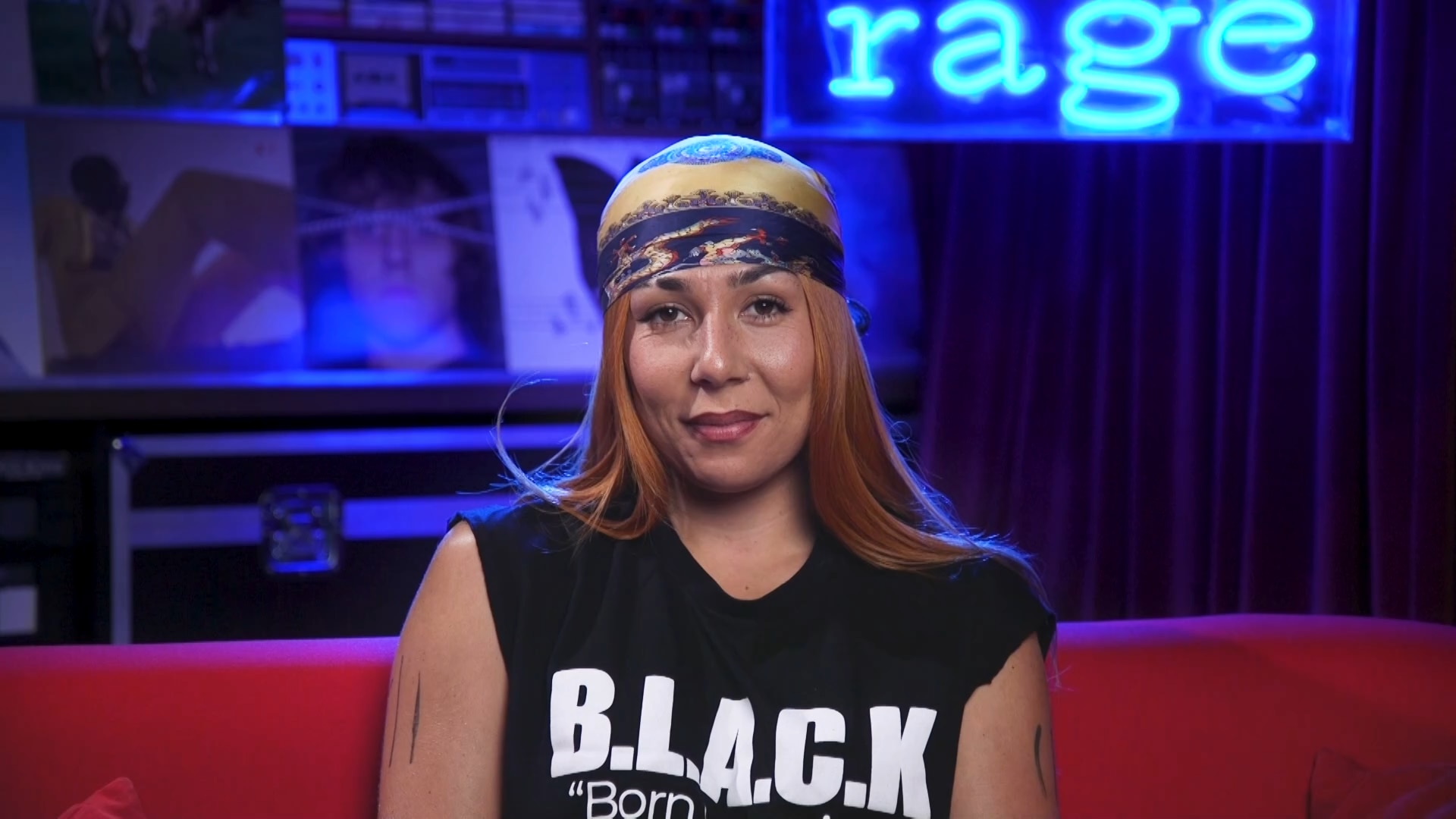 MIss Kaninna sits on a red couch in front of a neon blue sign that reads rage